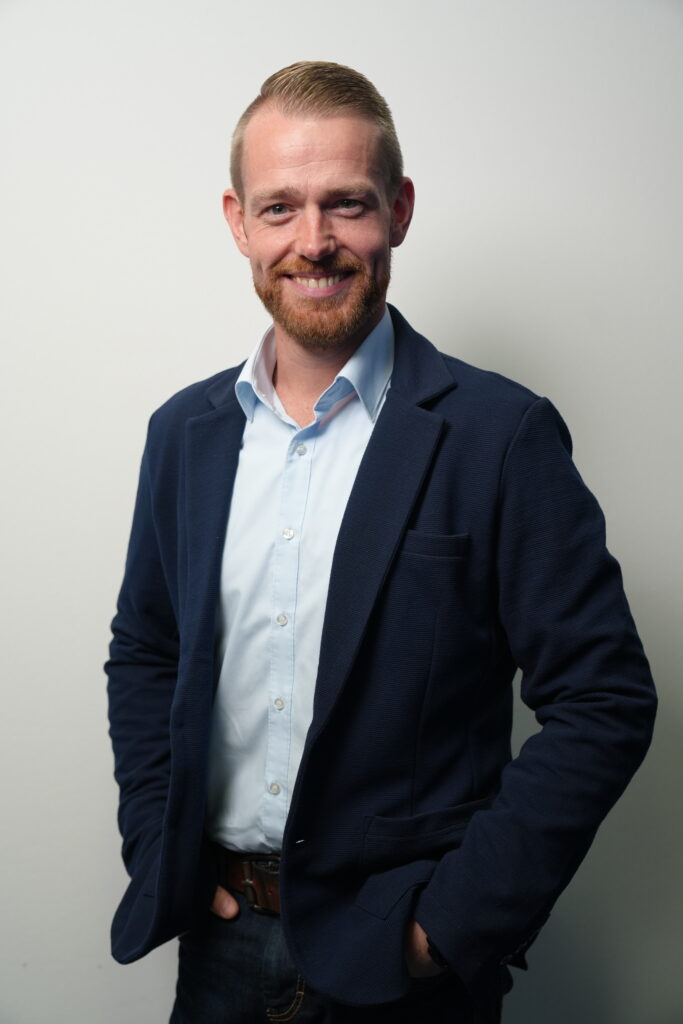 Ein lächelnder Mann mit Bart in dunkelblauem Sakko und hellblauem Hemd vor hellem Hintergrund